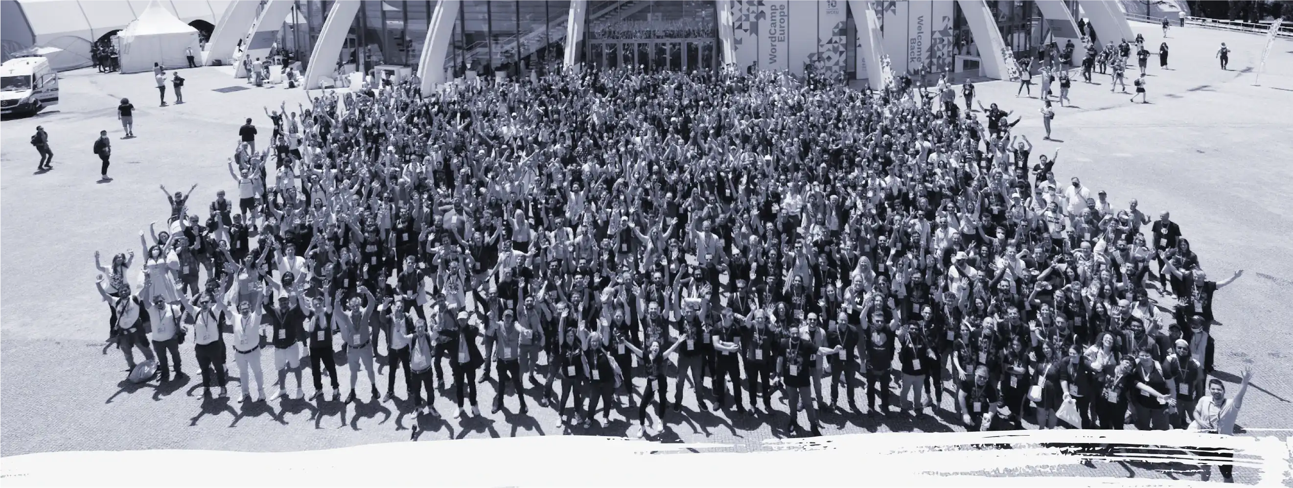 Una vista de pájaro en blanco y negro de los miles de colaboradores que asistieron a WordCamp Europa 2022. Todos ellos de pie frente al recinto con las manos en alto en señal de celebración. Foto: Nilo Vélez.