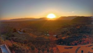 A panoramic view unveils a sunrise over a mountainous region. The sky transitions from a warm orange near the horizon to soft blue above. Sparse vegetation, undulating hills, and a glinting pond compose the serene landscape, with a lone building perched atop. Paths ribbon through the terrain.