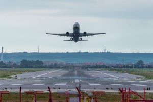 Front view of a Boeing 737-8AS airplane taking off from an airport runway, viewed from the perspective of the airport with a fence in the foreground and a hilly landscape with buildings in the background.