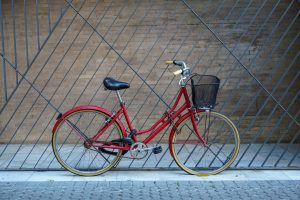 A red vintage bicycle with a basket parked against a modern geometric patterned fence on a stone-paved ground.
