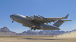 A C17 Globemaster military cargo plane takes off from a dusty airstrip, with rugged mountains in the background.