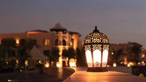 An ornamental gold lantern glowing with light outside on a balcony with a blurred town in the background.