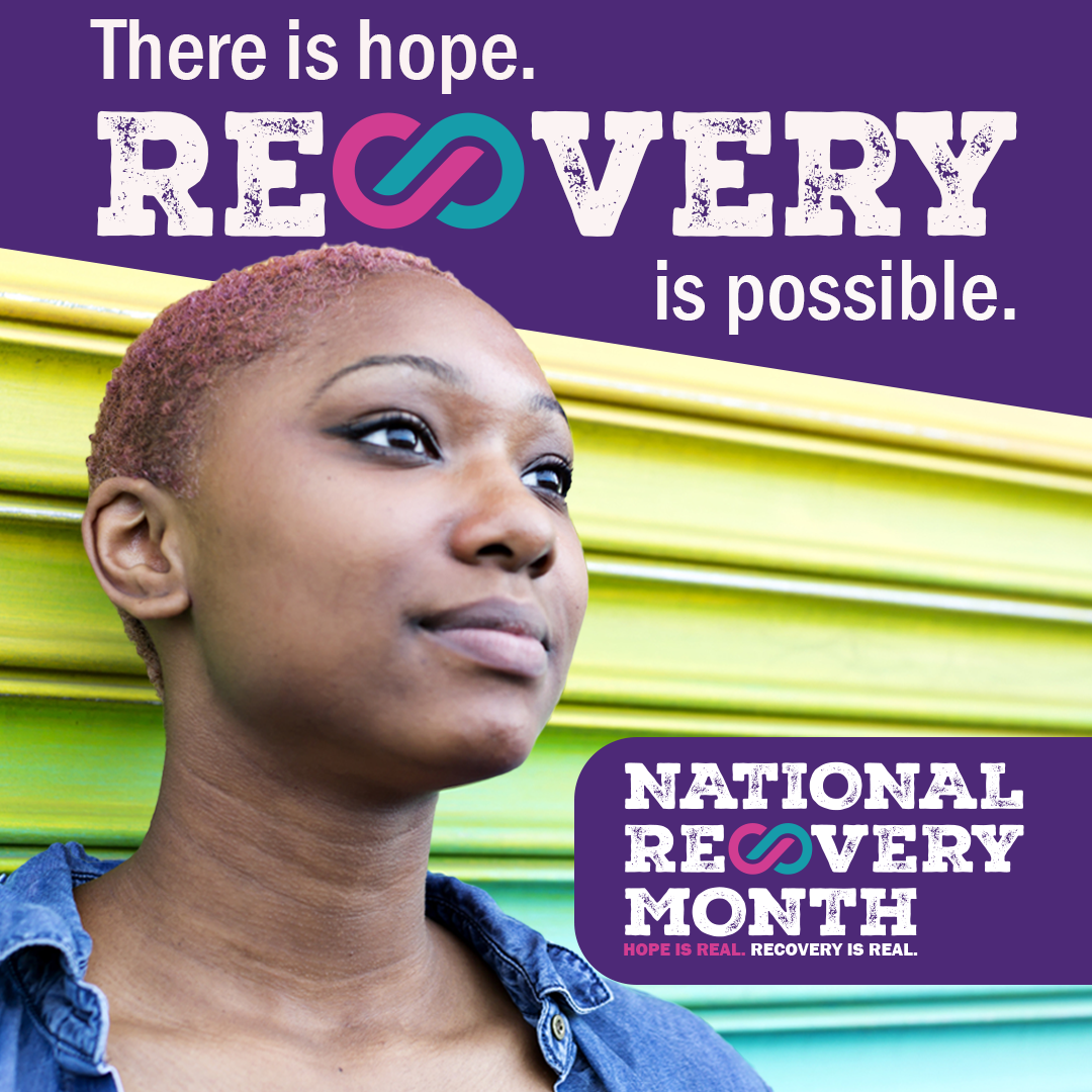 A person standing in front of a yellow, green, and blue wall with a banner that reads, “There is hope. Recovery is possible.” A logo in the bottom right corner reads, “National Recovery Month. Hope is Real. Recovery is Real.”