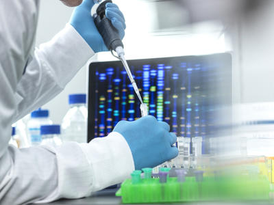 Close-up image of a scientist pipetting a sample into a vial for DNA testing. A screen showing DNA array results is in the background. 