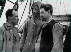 Three men in well-worn clothing stand on the deck of a ship. Man on right is thickset, bearded, with cigarette in mouth. Man in centre is hooded; man on left, also bearded and with receding hair, is shown right profile.