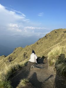 A trekker in a white jacket is sitting on a rocky path surrounded by grassy mountains under a blue sky with light clouds.