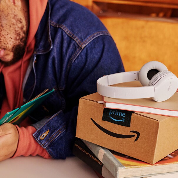 A man in a denim jacket opens a book to study; to his left on the table there's an unopened Amazon box, a mint green Takeya water bottle, and white Sony headphones.