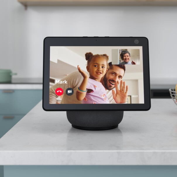 En una mesa de cocina está un dispositivo Echo realizando una vídeollamada. En la pantalla en grande sale Mark y su hija, los dos saludando. En la parte superior derecha está la mamá de Mark también saludando. La mamá lleva un jersey de cuello alto azul y el pelo rizado corto de color gris. Marc tiene barba y perilla y su hija es morena con el pelo rizado. La niña lleva tres pulseras en la mano derecha. En la mesa de la cocina hay un frutero con fruta yen el fondo, en la vitrocerámica una olla de color verde. La pared de la cocina es blanca. 