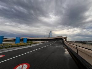 A rooftop racing track with fresh pavement and blue cylindrical structures on top of Lingotto building. The floor is wet from recent rain, with visible puddles along the edges.