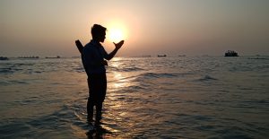 A serene seascape at sunset with a calm sea and gentle waves in the foreground. A man catching sunset, in the background there are many small boat and big ships.