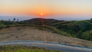 A scenic view of a sunset over rolling hills covered with vegetation. The sun is setting on the horizon, casting an orange and yellow glow across the sky. 