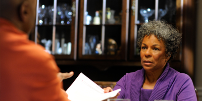 Older Black woman in a living room, showing a paper to a Black man sitting across the table.