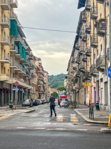 A man walks down a street in Torino, Italy. Buildings rise up either side of the street. Cars are parked on either side. The mountains can be seen in the distance.