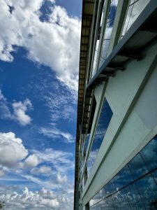 Looking up at a blue sky with white fluffy clouds beside a building reflecting the sky in its glass windows.