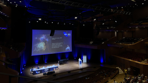 A dark conference room with high ceilings and a person presenting on stage. 