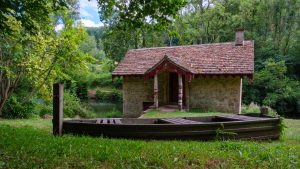 A wooden boat lies on the grass next to a post.  Behind the boat is an old boathouse with a lake just visible in the background.  The frame is dominated by the lush green trees.