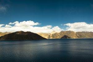 A calm lake with clear, blue water flanked by brown, rugged mountains under a clear blue sky with scattered white clouds