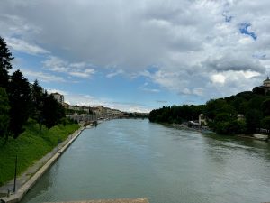 The photo captures a serene view of the Po River in Torino, Italy, taken just after crossing the Umberto Bridge. The river flows calmly, flanked by lush green trees on both sides. On the left bank, a walking path runs parallel to the river, lined with trees and lamp posts. Buildings and houses are visible in the distance, along with some small boat
