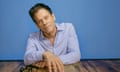 Kevin Bacon, in a pale blue shirt, arms and hands resting on a table next to his black-framed glasses