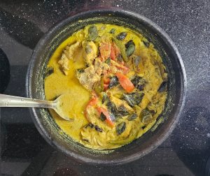 A traditional Kerala fish curry with tomatoes, and coconut milk, served in a black pot with a spoon, on a kitchen counter.
