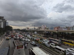 A very busy road in an Asian city, with numerous vehicles and an elevated railway. Several building can be seen in the background. Dark clouds cover the sky.