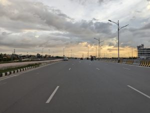 A four lane road with several vehicles, with a cloudy sky and beautiful orange sunlight in the background. Multiple buildings and street lights can be seen on either side of the road.