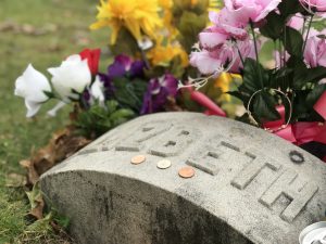 The gravestone of Lizzy Borden in Fall River, Massachusetts: a simple arched stone with Elizabeth carved onto the top. 3 coins are placed on top and colorful flowers are planted around. 