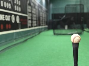 A batting cage with a baseball on a hitting tee in the foreground with an L screen and scoreboard out of focus in the background.