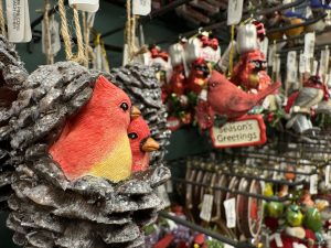 A Christmas ornament of a pine cone with two red cardinals nesting inside in the foreground and rows of other assorted ornaments in the background.