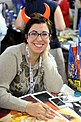 Die dunkelhaarige Zeichnerin Mirka Andolfo sitzt anlässlich einer Comic- und Spielemesse an einem Tisch; mit Brille und aufgesetzten Teufelshörnern schaut sie lächelnd in die Kamera.