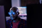 GLEAMING DATA IS REFLECTED IN THE EYGLASSES OF A MAN SEATED AT A COMPUTER, IN A DARKENED ROOM