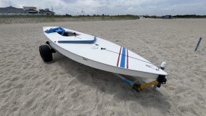 Single Sunfish sailboat sitting on a wheeled trailer in the sand with a blue life jacket on top and the Lewes Yacht Club in the background (Delaware, USA).