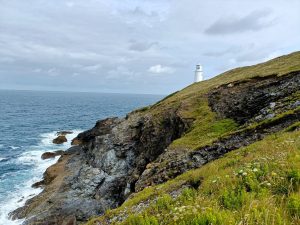 A white lighthouse perched on a grassy hill overlooking a rocky coastline with waves crashing against the rocks under a cloudy sky.