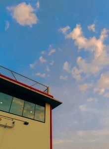 A modern building with large glass windows and red accents on the edge of the roof. Above is a bright blue sky with scattered, fluffy clouds.